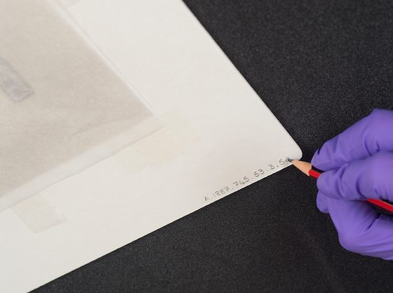 A gloved hand applying a label to a mounted artwork with a pencil.